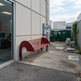 A red metal bench with a curved backrest is mounted against a light gray concrete building wall in an outdoor alleyway next to a glass door.