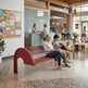 A red outdoor-style bench with curved supports is shown in a bright community center or library interior, with several people sitting and standing nearby. The scene includes a reception desk, tables, plants, and large windows.