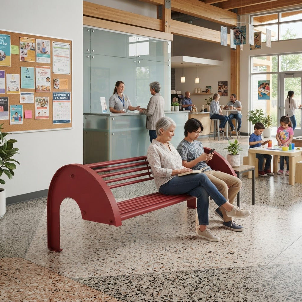 A red outdoor-style bench with curved supports is shown in a bright community center or library interior, with several people sitting and standing nearby. The scene includes a reception desk, tables, plants, and large windows.