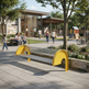 A modern outdoor park bench with bright yellow curved supports and silver metal slats, situated in a sunny urban plaza with people walking and sitting nearby.