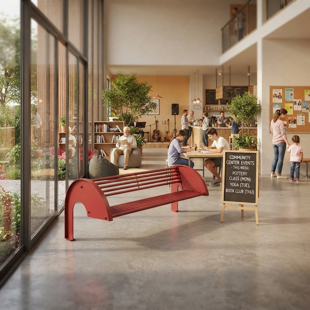 A red slatted outdoor-style bench with curved supports is placed in a bright modern community center lobby. Several people are seated and standing in the background near tables, plants, and a chalkboard sign.