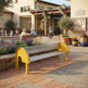 A modern outdoor bench with a bright yellow curved frame and silver metal slatted seat, placed on a paved plaza in front of a building with people sitting nearby.