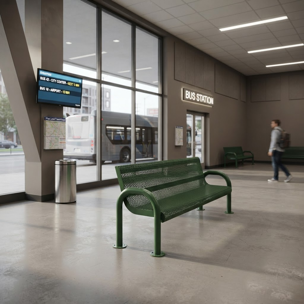 A green metal bench sits in a modern bus station interior with large windows, a bus outside, a map on a pole, and a person walking in the background.
