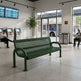 A green metal bench with a perforated mesh seat and back, placed in a modern transit station interior with large windows, ticketing kiosks, a bus outside, and seated people nearby.