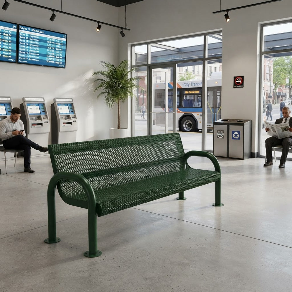 A green metal bench with a perforated mesh seat and back, placed in a modern transit station interior with large windows, ticketing kiosks, a bus outside, and seated people nearby.