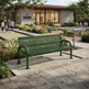 A green metal mesh park bench in the foreground of an outdoor plaza, with a modern building, landscaped plants, and a few people walking in the background.