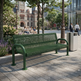 A green metal mesh park bench in an outdoor urban plaza, shown with a trash can, trees, and a historic stone building in the background.