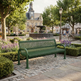 A green metal mesh park bench situated in a beautifully landscaped town square with a fountain, historic building, and trees in the background.