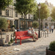 A bright red metal park bench in a sunlit urban plaza, with two elderly people sitting on it. Trees, buildings, pedestrians, a lamppost, and storefronts are visible in the background.