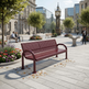 A maroon metal bench with a perforated slatted seat and backrest, positioned in a modern urban plaza with stone paving, trees, and buildings in the background.