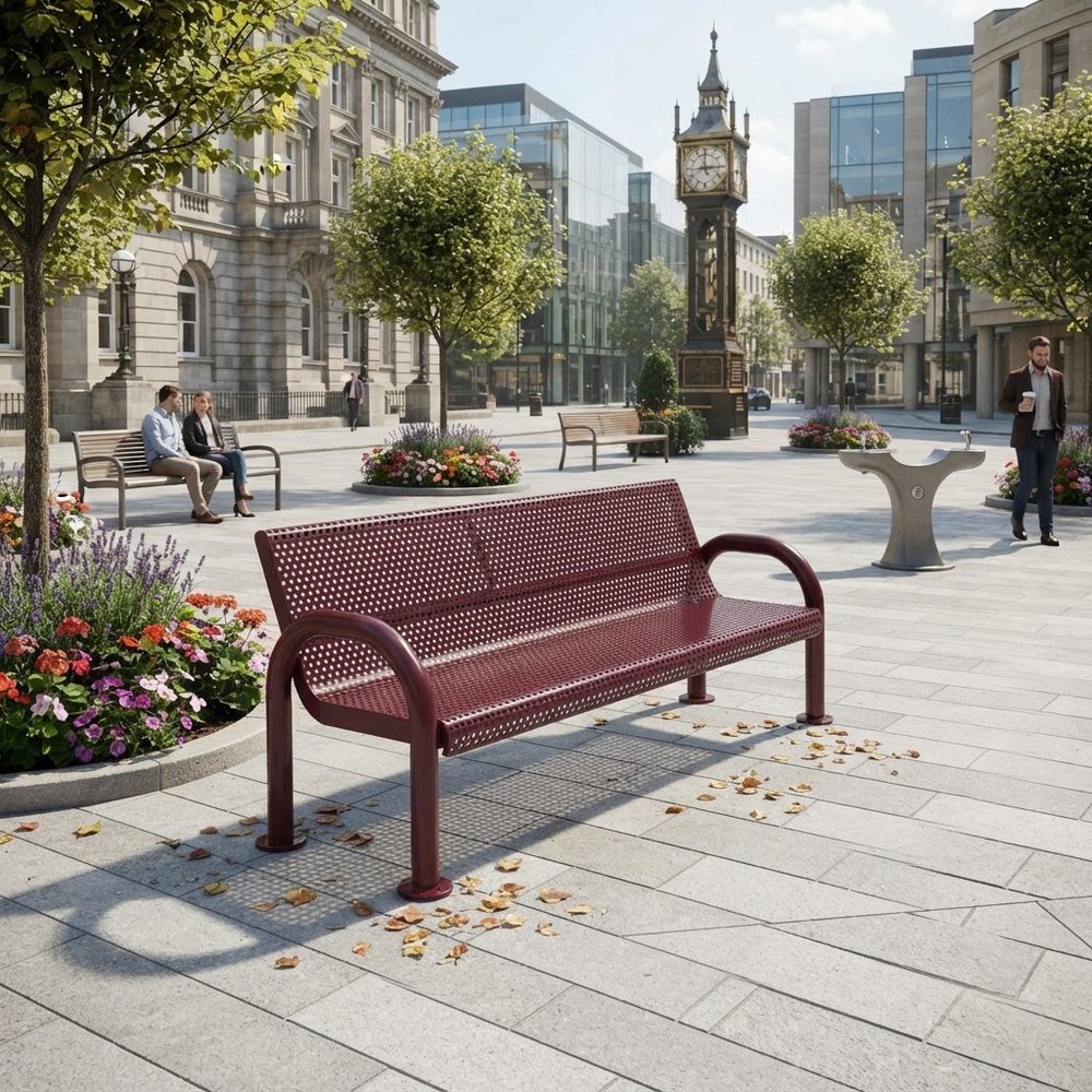 A maroon metal bench with a perforated slatted seat and backrest, positioned in a modern urban plaza with stone paving, trees, and buildings in the background.