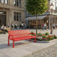 A bright red metal bench with a perforated mesh design is situated in a city plaza. The scene includes a stone pavement, a cobblestone section, a small flower bed, a tree, and a classic street lamp.