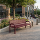 A maroon metal bench with a slotted backrest sits on a paved plaza in front of a modern building, with trees, flower beds, and a small informational sign nearby.