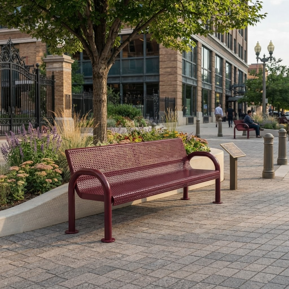 A maroon metal bench with a slotted backrest sits on a paved plaza in front of a modern building, with trees, flower beds, and a small informational sign nearby.