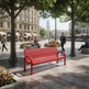 A bright red perforated metal bench in a modern city plaza, surrounded by trees, pedestrians, and buildings.
