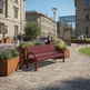 A red metal park bench with a perforated mesh seat and back, situated outdoors in a stone plaza with people, plants, and historic buildings in the background.