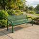A green metal park bench with curved armrests sits on a concrete path in a landscaped garden with trees, flowers, and a pathway leading into the background.