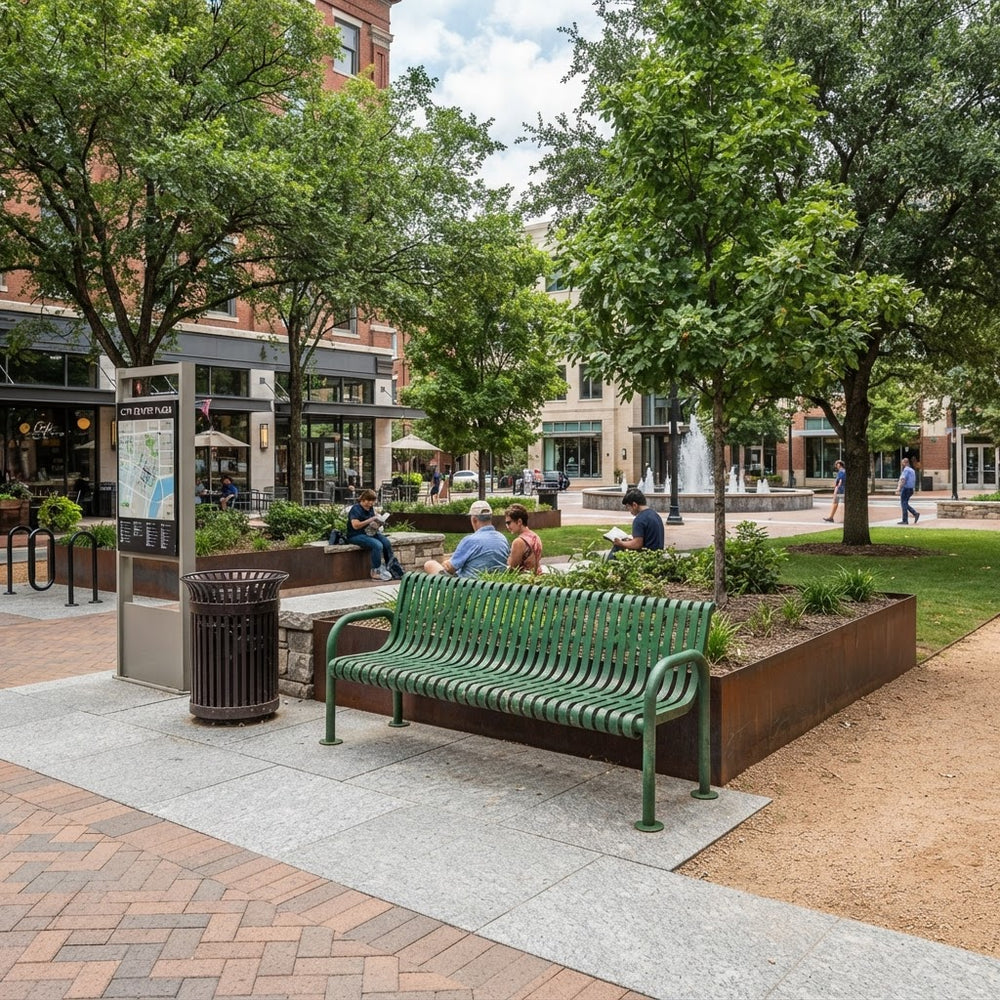 A green metal park bench sits on a paved plaza in a city square, with trees, grass, people sitting nearby, and storefront buildings in the background.