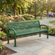 A green metal park bench with curved armrests sits on a stone patio in a sunny garden park. Flower beds, trees, and a walking path with distant people are visible in the background.