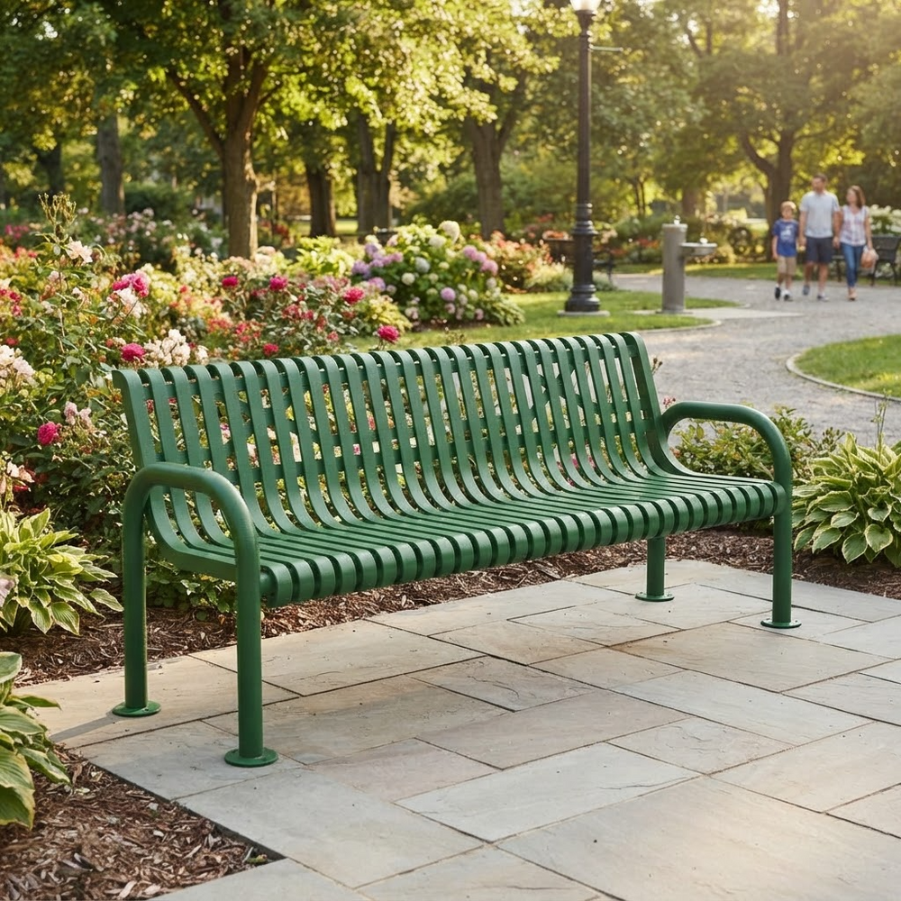 A green metal park bench with curved armrests sits on a stone patio in a sunny garden park. Flower beds, trees, and a walking path with distant people are visible in the background.