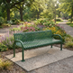 A green metal park bench with curved armrests sits on a concrete pad in a landscaped garden with flower beds and a tree-lined path in the background.