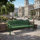A green slatted outdoor bench in a paved public square with trees, people, and a large classical building in the background.