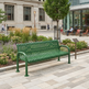 A green metal park bench with curved armrests and slatted back and seat, situated on a paved plaza in an urban outdoor setting with trees, flowers, and nearby seating areas.