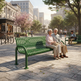 A green metal park bench in a city plaza, with an elderly couple sitting on it. The scene is set in a sunny urban park with trees, other benches, pedestrians, and buildings in the background.