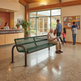 A green metal bench with a dark frame sits in a bright community center lobby. A man sits on the bench reading, while other people walk near large windows and a reception desk in the background.