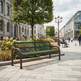 A green metal park bench with dark brown armrests sits on a paved plaza in a city square, surrounded by flower beds, trees, and pedestrians walking between modern and historic buildings.