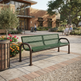 A green metal mesh park bench with dark brown curved armrests, situated on a paved plaza in front of a brick community center building with trees and people in the background.