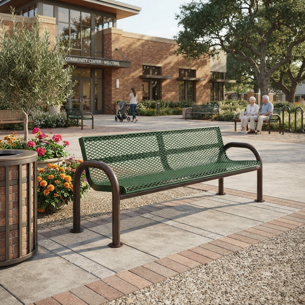 A green metal mesh park bench with dark brown curved armrests, situated on a paved plaza in front of a brick community center building with trees and people in the background.