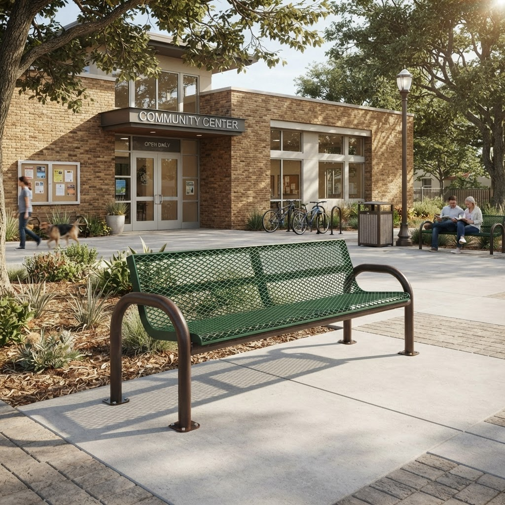 A green metal mesh park bench with dark brown curved armrests and legs, situated on a concrete plaza in front of a brick community center building with trees and people in the background.