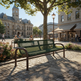 A green metal park bench in a sunny town square with stone paving, a large tree, and historic stone buildings in the background. A few people are seated at tables nearby.