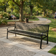 A black metal park bench with a slatted back and seat, placed on a stone path in a sunny, tree-lined park. In the background, people are sitting on a bench and a dog is walking near a trash can.