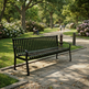 A black metal park bench with a slatted back and seat, positioned on a stone pathway in a lush green park with trees, flowers, and distant people.