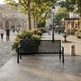 A black metal park bench with a slatted back and seat, placed on a paved plaza in a city square with trees, pedestrians, and buildings in the background.