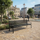 A black metal slat bench in a modern town square with paved stone walkways, trees, benches, a lamppost, and distant buildings under a blue sky.