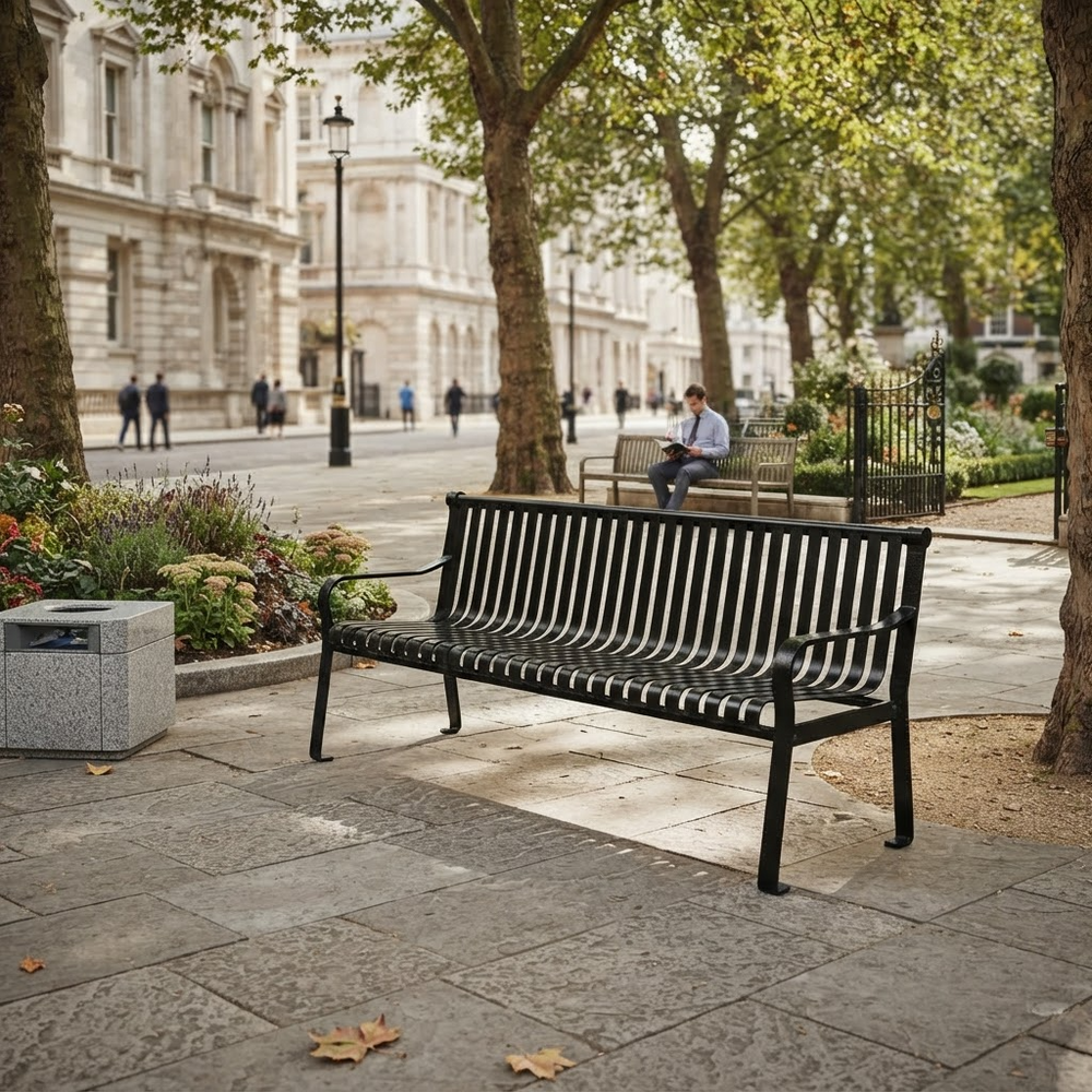 A black metal park bench with a slatted back and seat, placed on a stone paved walkway in a city park. Trees, buildings, and a seated person are visible in the background.