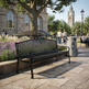 A black metal park bench with a slatted back and seat, shown outdoors on a stone plaza beside a tree, flowers, and a trash can. Pedestrians and a historic building are visible in the background.