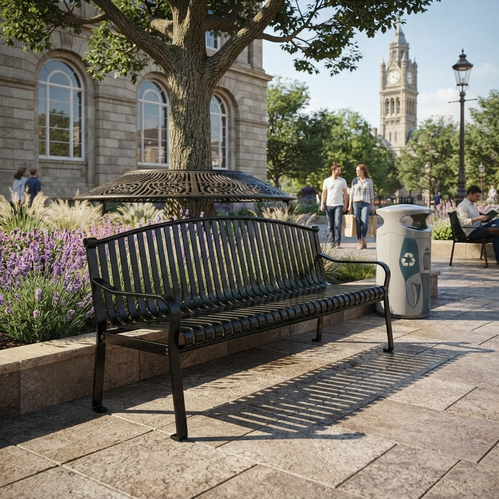 A black metal park bench with a slatted back and seat, shown outdoors on a stone plaza beside a tree, flowers, and a trash can. Pedestrians and a historic building are visible in the background.