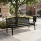 A black metal park bench with a slatted back and seat, placed outdoors on a paved plaza beside a tree and city buildings. A dark trash can is at the right side of the bench.
