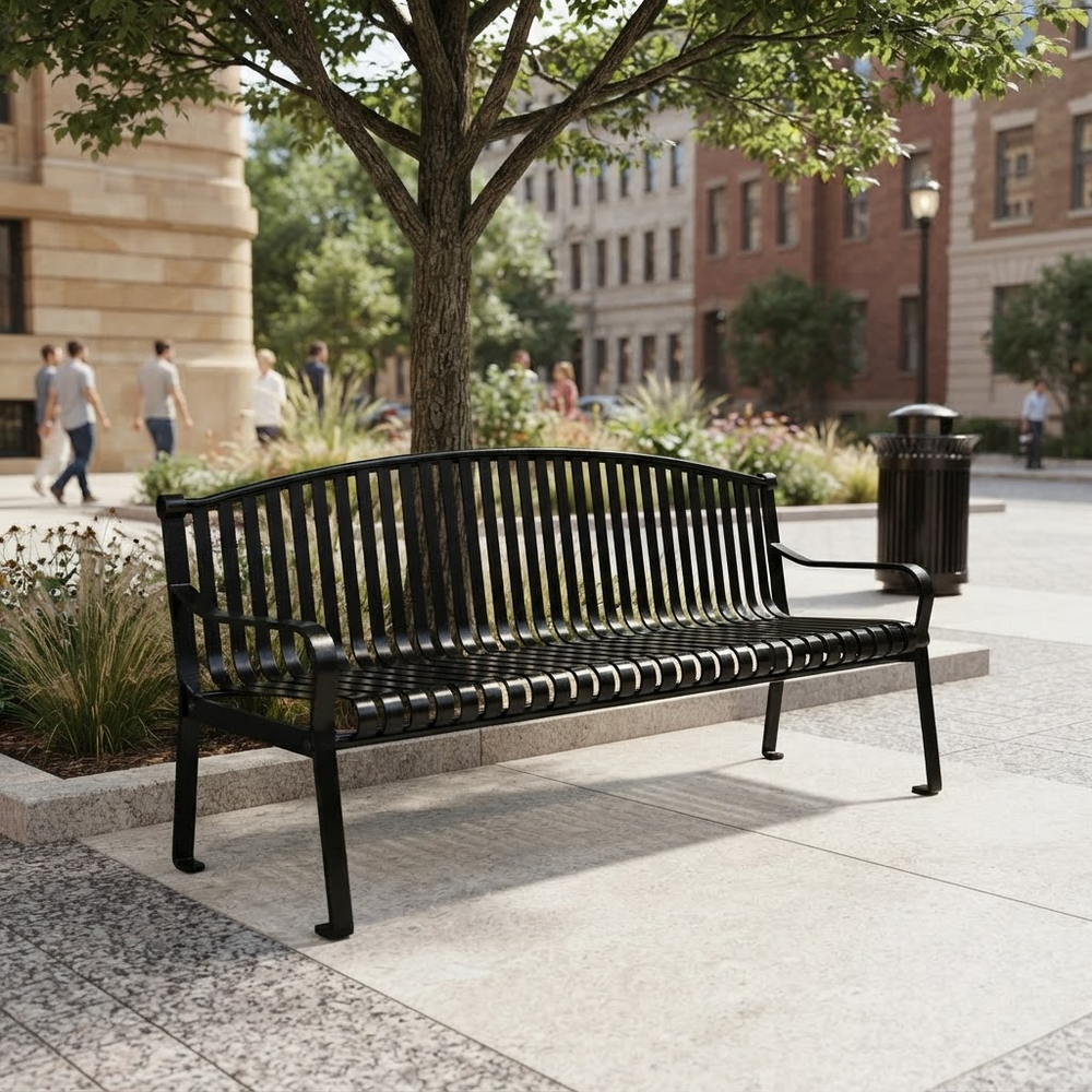 A black metal park bench with a slatted back and seat, placed outdoors on a paved plaza beside a tree and city buildings. A dark trash can is at the right side of the bench.