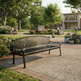 A black metal park bench with a slatted back and seat, placed on a paved walkway in a landscaped outdoor plaza with trees, people, and a building in the background.