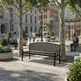 A black metal park bench is shown in a real outdoor plaza with trees, people sitting nearby, and city buildings in the background.