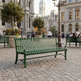 A green metal park bench in the foreground of a city plaza with stone paving, large classical buildings, trees, and blurred pedestrians in the background.