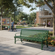 A green slatted outdoor bench in the foreground of a sunny park scene with people walking near a public recreation center, trees, flowers, and playground equipment in the background.