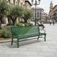 A green slatted metal park bench in a paved public square, with a man sitting on the bench reading a newspaper and historic buildings in the background.