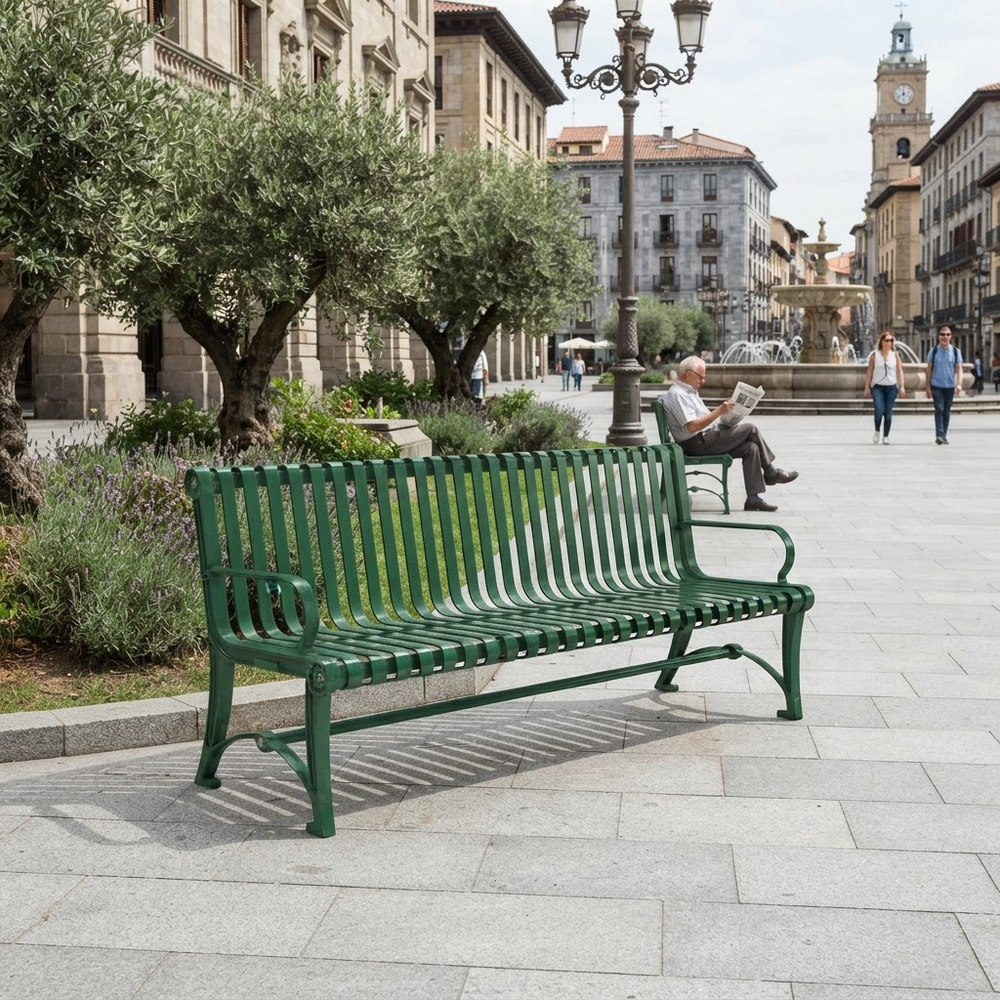 A green slatted metal park bench in a paved public square, with a man sitting on the bench reading a newspaper and historic buildings in the background.