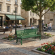 A green metal park bench is shown in a real outdoor plaza with stone paving, a tree, flower beds, and people walking and sitting nearby. Because a real environment and people are present, this is Lifestyle rather than Product/Packaging.